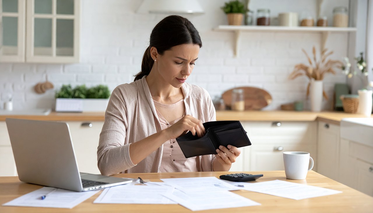 Eine junge Frau bei der Finanzplanung in der Küche: Laptop, Taschenrechner, Papiere und Geldbeutel auf dem Tisch zur Haushaltsbudget-Überprüfung.