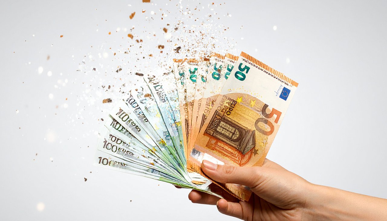 Hand hält Euro-Banknoten, die in kleine Partikel zerfallen – Symbolbild für Geldverlust durch Inflation und steigende Schulden.