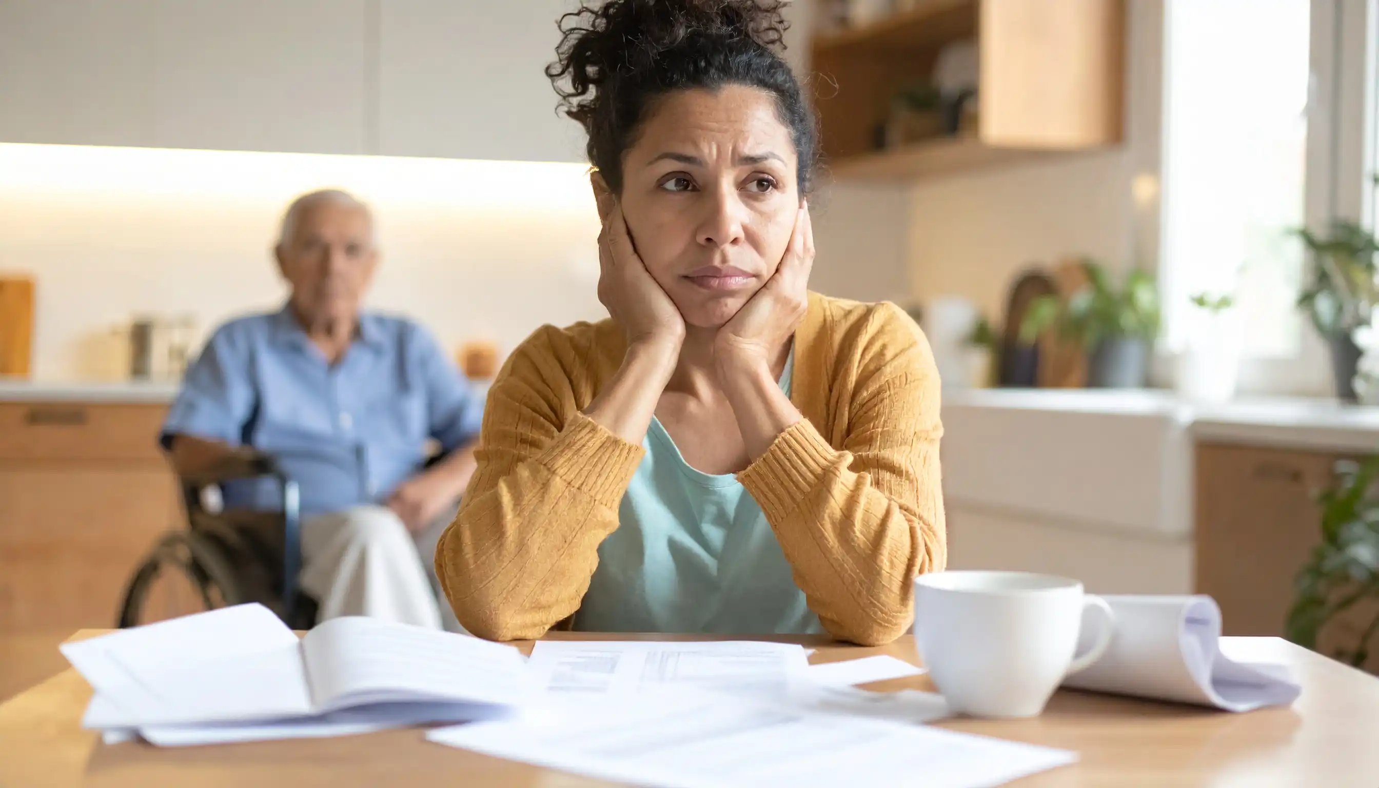 Eine Person mittleren Alters sitzt besorgt am Küchentisch mit Rechnungen und Pflegeunterlagen, im Hintergrund unscharf eine ältere Person im Rollstuhl.