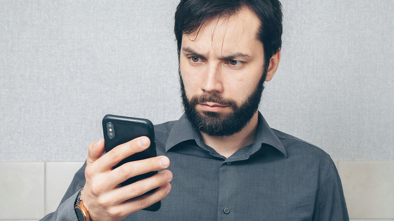 Sind Mahnungen per Mail oder WhatsApp zulässig? Skeptischer Mann schaut auf sein Smartphone